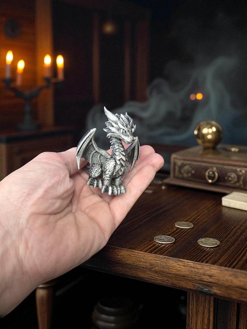 Puede incluir: Una detallada figura de drag&oacute;n plateado con alas extendidas, sostenida en una mano. El drag&oacute;n tiene escamas intrincadas y una cresta puntiaguda. El fondo incluye una mesa de madera, monedas y un entorno borroso y atmosf&eacute;rico con velas.