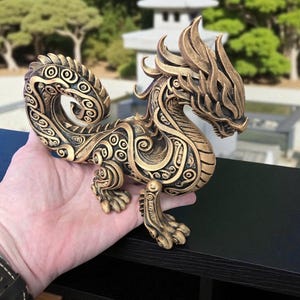 May include: A detailed bronze-colored dragon figurine with intricate patterns and a stylized design. The dragon has a long, curved tail and is posed on all fours. The background features a blurred outdoor scene with trees and a pagoda.
