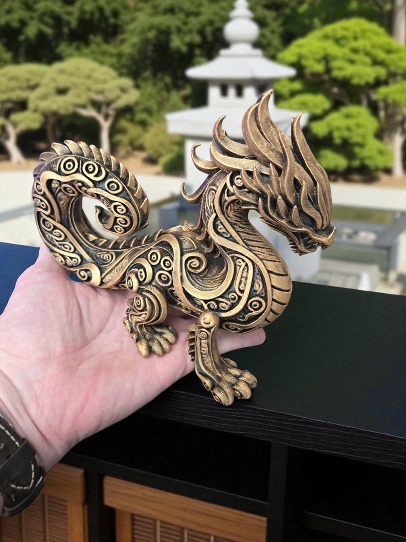 May include: A detailed bronze-colored dragon figurine with intricate patterns and a stylized design. The dragon has a long, curved tail and is posed on all fours. The background features a blurred outdoor scene with trees and a pagoda.