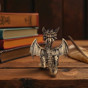Puede incluir: Una figura de drag&oacute;n de color bronce con alas y escamas detalladas sobre una superficie de madera. Una pluma y libros apilados est&aacute;n en el fondo. La figura tiene un acabado texturizado y est&aacute; bien iluminada.