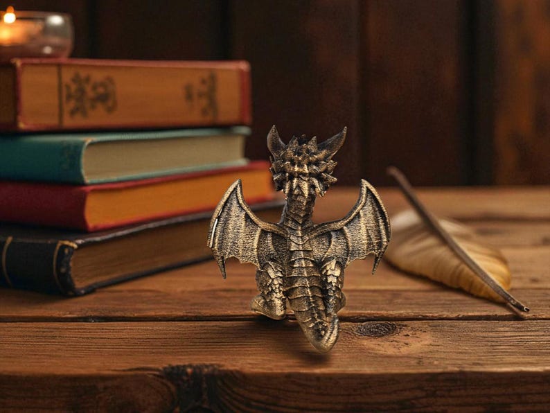Puede incluir: Una figura de drag&oacute;n de color bronce con alas y escamas detalladas sobre una superficie de madera. Una pluma y libros apilados est&aacute;n en el fondo. La figura tiene un acabado texturizado y est&aacute; bien iluminada.