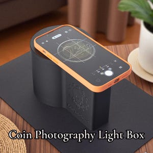 May include: A black coin photography light box with a smartphone resting on top. The phone has an orange case and is displaying a coin image. The text "Coin Photography Light Box" is at the bottom of the image.