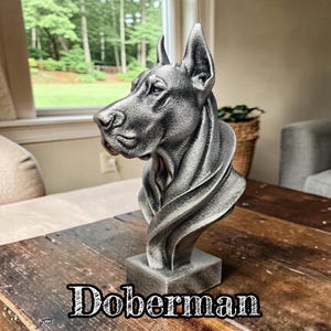 May include: A silver-toned Doberman dog bust sculpture featuring a detailed head and neck profile with pointed ears, set on a rectangular base. The surface has a textured, metallic finish. The word Doberman is printed in white text.