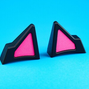 May include: Two black cat ear accessories with bright pink triangular inserts, set against a vibrant blue background. The cat ears have a geometric design, perfect for cosplay or adding a playful touch to any outfit.