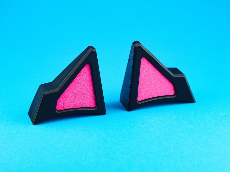 May include: Two black cat ear accessories with bright pink triangular inserts, set against a vibrant blue background. The cat ears have a geometric design, perfect for cosplay or adding a playful touch to any outfit.