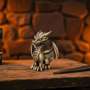 Puede incluir: Una detallada figura de drag&oacute;n de color bronce con escamas, alas y cuernos intrincados. El drag&oacute;n est&aacute; posado sobre una superficie de madera, con un fondo oscuro iluminado por velas. Una varita decorativa se encuentra cerca.