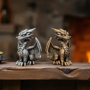 Puede incluir: Dos figuras detalladas de dragones, una plateada y otra dorada, se colocan una al lado de la otra sobre una superficie de madera. Los dragones tienen escamas intrincadas, alas y una expresi&oacute;n feroz. El fondo est&aacute; borroso, con una chimenea c&aacute;lida visible.