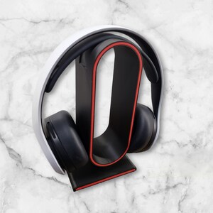 May include: A black headphone stand with a red accent, designed to hold a pair of white headphones.