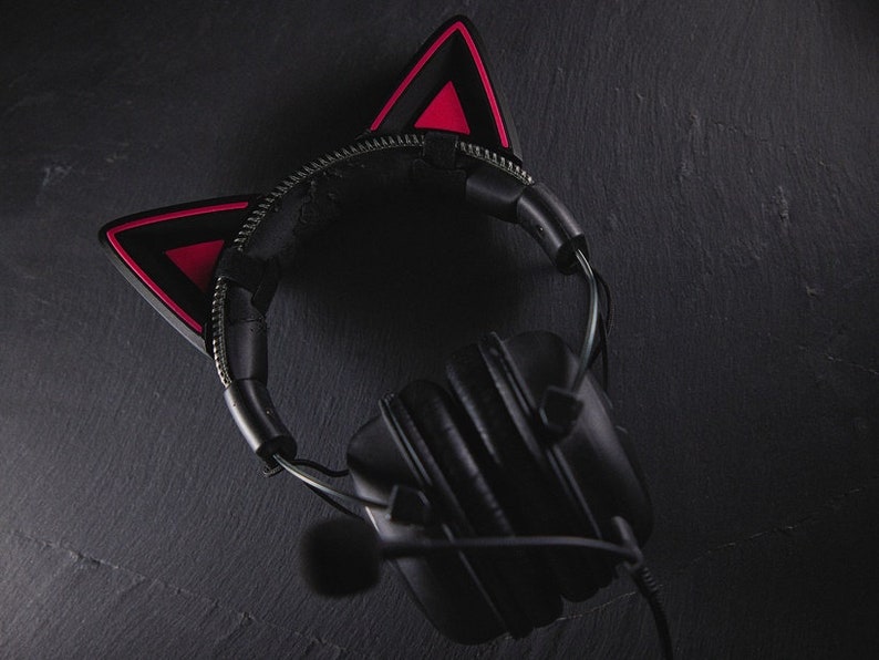Puede incluir: Auriculares negros con diadema negra y orejas de gato rojas. Los auriculares tienen un micr&oacute;fono conectado al auricular izquierdo.