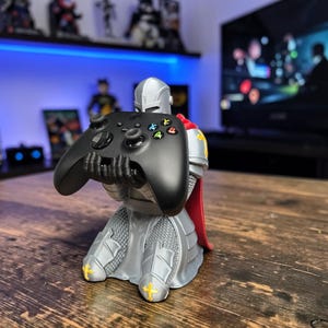 May include: A gray knight figurine kneeling and holding a black wireless game controller. The knight wears silver-toned armor with a red cape and features small yellow cross emblems on the knee guards. The controller has black buttons.