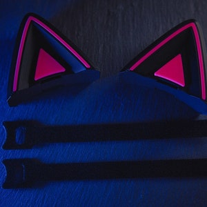 Puede incluir: Dos auriculares negros con forma de orejas de gato con detalles rosas. Los auriculares est&aacute;n sobre una superficie azul oscuro con dos correas negras.