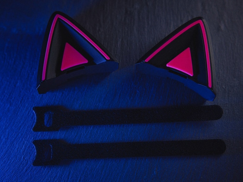 Puede incluir: Dos auriculares negros con forma de orejas de gato con detalles rosas. Los auriculares est&aacute;n sobre una superficie azul oscuro con dos correas negras.