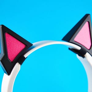 May include: White headphones with black and pink cat ears. The cat ears are black with pink triangular inserts. The headphones have a white band and are set against a bright blue background. The design is modern and playful.