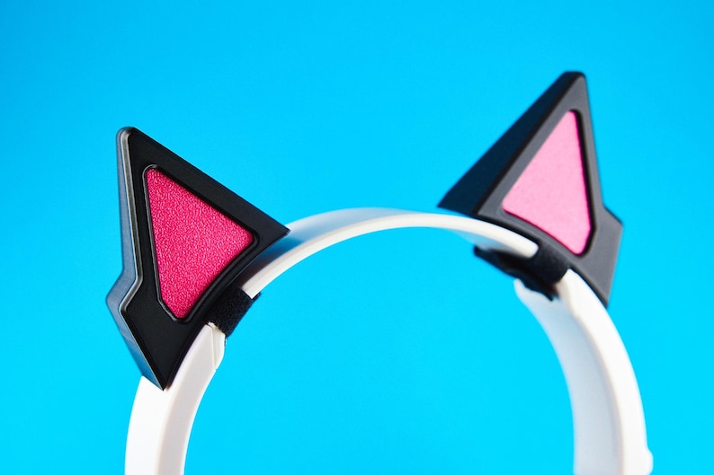 May include: White headphones with black and pink cat ears. The cat ears are black with pink triangular inserts. The headphones have a white band and are set against a bright blue background. The design is modern and playful.