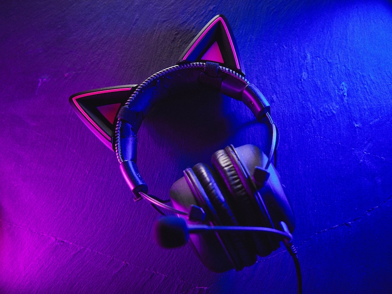 Puede incluir: Auriculares de juego negros con orejas de gato rosas, sobre un fondo azul y morado.