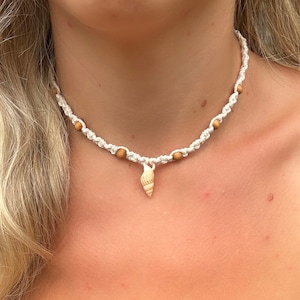 Pode incluir: Um colar gargantilha de macramé com um pequeno pingente de concha. O colar é feito de cordão branco sujo, com pequenas contas de madeira espaçadas ao longo do fio. A concha é bege claro com listras marrons.
