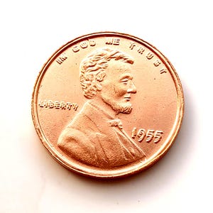 May include: A copper-colored United States penny coin. The obverse features a profile of Abraham Lincoln, the word "LIBERTY", and the year "1955". The coin also has the inscription "IN GOD WE TRUST".