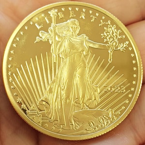 May include: A gold coin with a detailed depiction of Lady Liberty, holding a torch and an olive branch. The word "LIBERTY" is inscribed at the top, and the year "2023" is at the bottom. The coin has a bright, reflective surface.