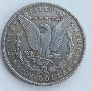 May include: A silver one-dollar coin with an eagle design, the words "United States of America" and "In God We Trust" are visible. The coin also features the words "One Dollar."
