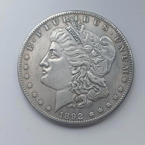 Op de afbeelding: Een zilveren munt met een gedetailleerd profiel van een vrouw met een kroon, het woord "LIBERTY" is zichtbaar. De rand van de munt is versierd met sterren en het jaartal "1892" is gegraveerd. De woorden "E PLURIBUS UNUM" zijn ook zichtbaar.