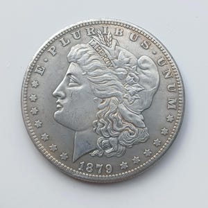 May include: A silver coin with a profile of a woman, the words "E PLURIBUS UNUM" and stars around the edge. The coin also features the word "LIBERTY" and the year 1879.