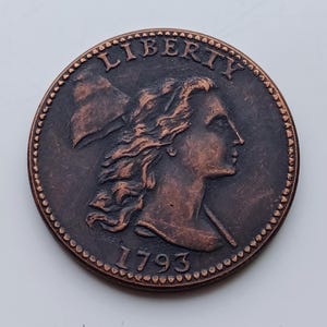 May include: A copper-colored coin featuring a profile of a woman with flowing hair, the word "LIBERTY" arched above, and the date "1793" below. The coin has a detailed edge and a slightly worn appearance.