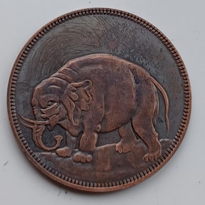 May include: A circular copper coin with a detailed relief of an elephant. The elephant is shown in profile, with tusks and a textured hide. The coin has a textured edge and a slightly aged appearance, suggesting it is an antique or vintage piece.