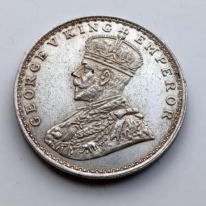 Puede incluir: Una moneda de plata con el perfil coronado de un hombre, con la inscripción "GEORGE V KING EMPEROR" alrededor del borde. La moneda presenta un diseño detallado y signos de desgaste, lo que indica su importancia histórica.