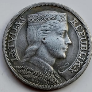 Puede incluir: Una moneda de plata con el perfil de una mujer que lleva un tocado decorativo. El borde de la moneda tiene un patrón texturizado. Las palabras "LATVIJAS" y "REPUBLIKA" están inscritas alrededor del perímetro de la moneda.