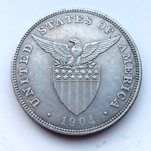 May include: A silver-toned United States of America coin dated 1904. The obverse features an eagle with outstretched wings perched above a shield containing stars and vertical stripes. The text UNITED STATES OF AMERICA is inscribed along the rim.