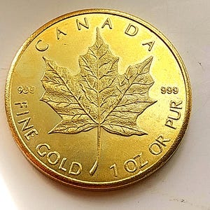 May include: A circular gold coin featuring a detailed maple leaf at the center. The text CANADA is arched at the top, with 999 on both sides of the leaf. The bottom text reads FINE GOLD 1 OZ OR PUR, indicating the weight is 1 ounce.