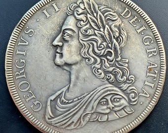 May include: A silver coin featuring a profile of a man with a laurel wreath and flowing hair. The coin has the inscription "GEORGIVS II DEI GRATIA" around the edge. The coin's rim has a detailed, ridged pattern.