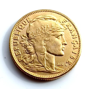 May include: A gold coin with a detailed profile of a woman wearing a laurel wreath. The coin's edge is textured, and the words "REPUBLIQUE FRANÇAISE" are inscribed around the perimeter. The coin has a shiny, reflective surface.