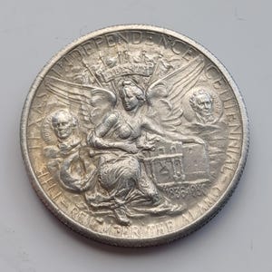 May include: A silver coin with intricate details. The coin features a central seated figure with wings, surrounded by text that reads "INDEPENDENCE CENTENNIAL" and "REMEMBER THE ALAMO". The coin also includes the years "1836-1936".