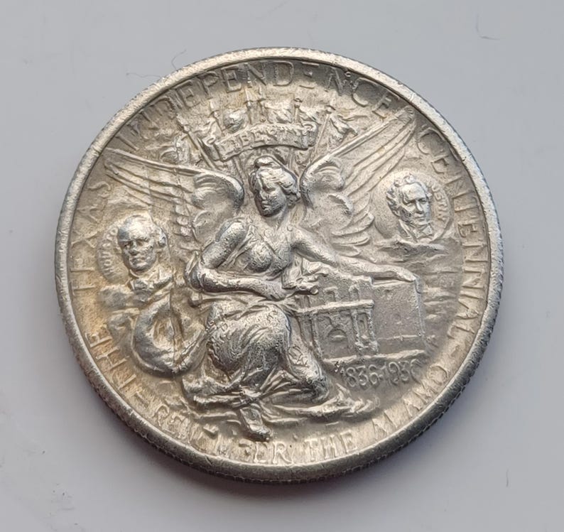 May include: A silver coin with intricate details. The coin features a central seated figure with wings, surrounded by text that reads "INDEPENDENCE CENTENNIAL" and "REMEMBER THE ALAMO". The coin also includes the years "1836-1936".