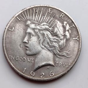 Könnte beinhalten: Eine silberne Peace-Dollar-Münze von 1926. Die Vorderseite zeigt ein Profil von Lady Liberty mit Strahlen, die von ihrer Krone ausgehen, sowie die Inschriften "IN GOD WE TRUST" und "LIBERTY". Das Prägejahr "1926" ist ebenfalls sichtbar.