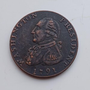 Może przedstawiać: Miedziana moneta z profilem George'a Washingtona. Moneta jest oznaczona napisem "WASHINGTON PRESIDENT" i rokiem "1791". Moneta ma szczegółowy portret i jest prawdopodobnie przedmiotem kolekcjonerskim lub historycznym.