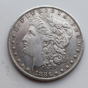 May include: A silver coin with a detailed portrait of a woman in profile, surrounded by text and stars. The coin is dated 1886. The words "E PLURIBUS UNUM" and "LIBERTY" are visible.