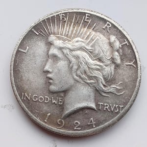 May include: A silver 1924 Peace dollar coin. The obverse features a profile of Lady Liberty with rays emanating from her crown, the word "LIBERTY" and the motto "IN GOD WE TRUST". The date "1924" is visible.