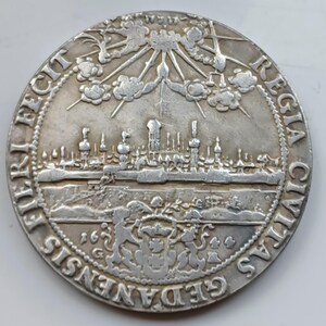 Puede incluir: Una moneda de plata con una detallada vista de una ciudad con torres y un sol radiante. La moneda tiene inscripciones en un patrón circular alrededor del borde, con el año 1659 visible. El diseño incluye nubes y un escudo de armas.