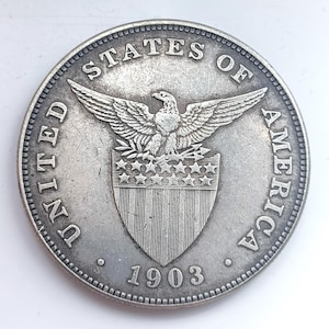 May include: A silver coin from 1903. The coin features an eagle with outstretched wings above a shield with stars and stripes. The coin is inscribed with "UNITED STATES OF AMERICA" around the edge.