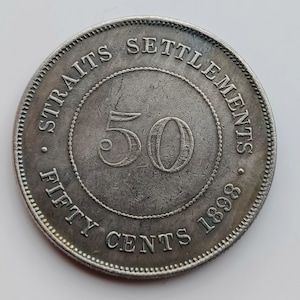 May include: A silver Straits Settlements 50 Cents coin from 1898. The coin is inscribed with "STRAITS SETTLEMENTS" at the top, "FIFTY CENTS" at the bottom, and the year "1898" on the right. The number "50" is in the center.