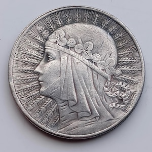 Puede incluir: Moneda de plata con el perfil de una mujer, adornada con una corona floral y cubierta con un velo. El borde de la moneda está detallado con espigas de trigo y una serpiente estilizada. La moneda tiene una apariencia antigua y detallada.