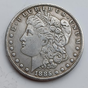 Puede incluir: Una moneda de plata Morgan de 1885. La moneda presenta un perfil de Lady Liberty, las palabras "E PLURIBUS UNUM" y "LIBERTY", y la fecha "1885". La moneda tiene un diseño detallado y una apariencia desgastada.