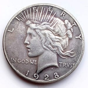 May include: A silver Peace dollar coin from 1928. The coin features a profile of Lady Liberty with the inscription "IN GOD WE TRUST." The reverse side of the coin is not visible. The coin is round and has a detailed edge.
