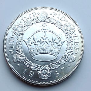 May include: A silver coin featuring a detailed crown design at the center, surrounded by floral and thistle motifs. The coin has text around the edge, including the year 1937. The coin has a detailed edge.