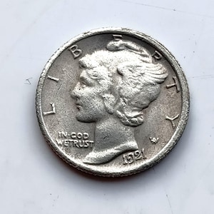 May include: A silver Mercury dime from 1921, featuring a profile of a woman with winged hair. The coin has the inscription "IN GOD WE TRUST" and the year "1921". The coin is a collectible item.