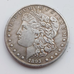 Puede incluir: Una moneda de plata Morgan de 1893. La moneda presenta el perfil de una mujer con cabello suelto, una corona y la palabra "LIBERTY". El borde de la moneda está detallado con estrellas y las palabras "E PLURIBUS UNUM."