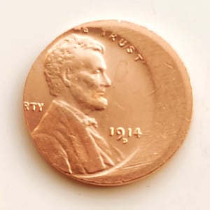 May include: A copper 1914 Lincoln penny. The obverse side shows a profile of Abraham Lincoln, with the words "LIBERTY" and "IN GOD WE TRUST". The year "1914" is also displayed on the coin.