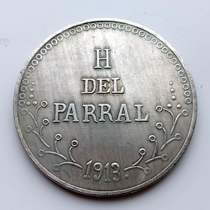 Pode incluir: Uma moeda de prata com a inscrição "H DEL PARRAL" e o ano "1913". A moeda tem uma borda decorativa com pequenos círculos e desenhos florais. A moeda parece antiga e desgastada.